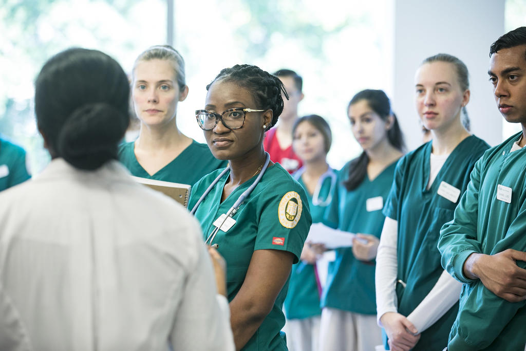Group of nursing students in green scrubs gather around instructor in white coat.