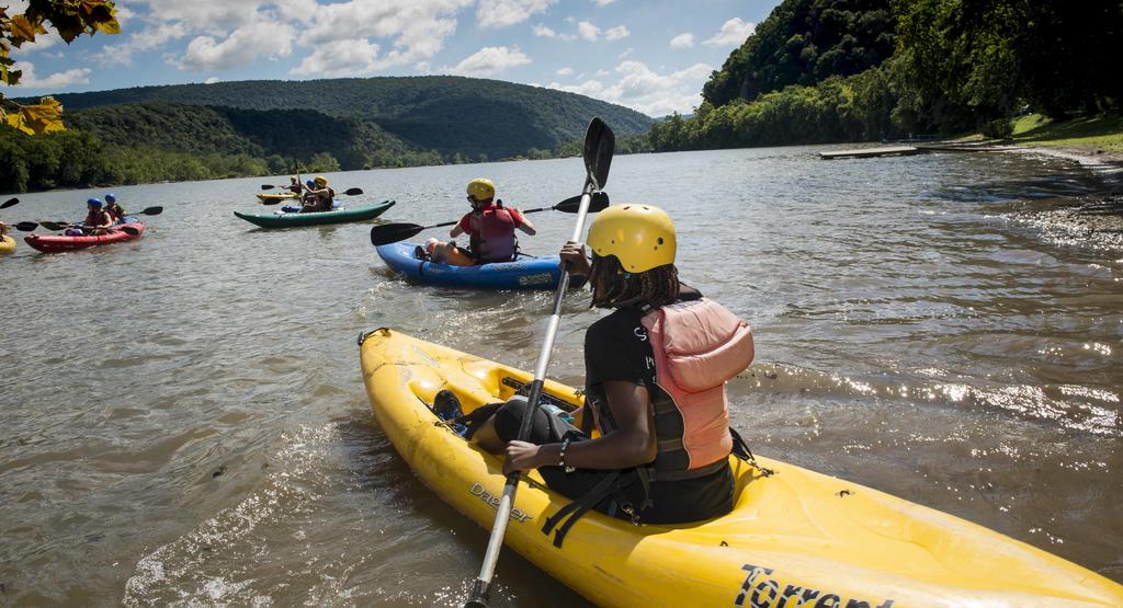 Group of kayakers wearing helmets and life jackets paddle along a river.