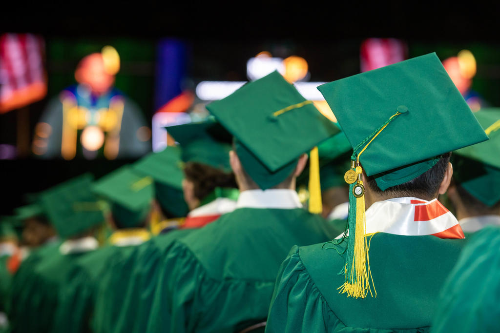 Students sit in rows wearing green caps and gowns for graduation at Mason's Patriot Center.