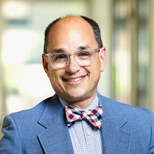 Smiling man wearing glasses and blue suit with red, white, and blue plaid bow tie.