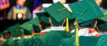 Students sit in rows wearing green caps and gowns for graduation at Mason's Patriot Center. 
